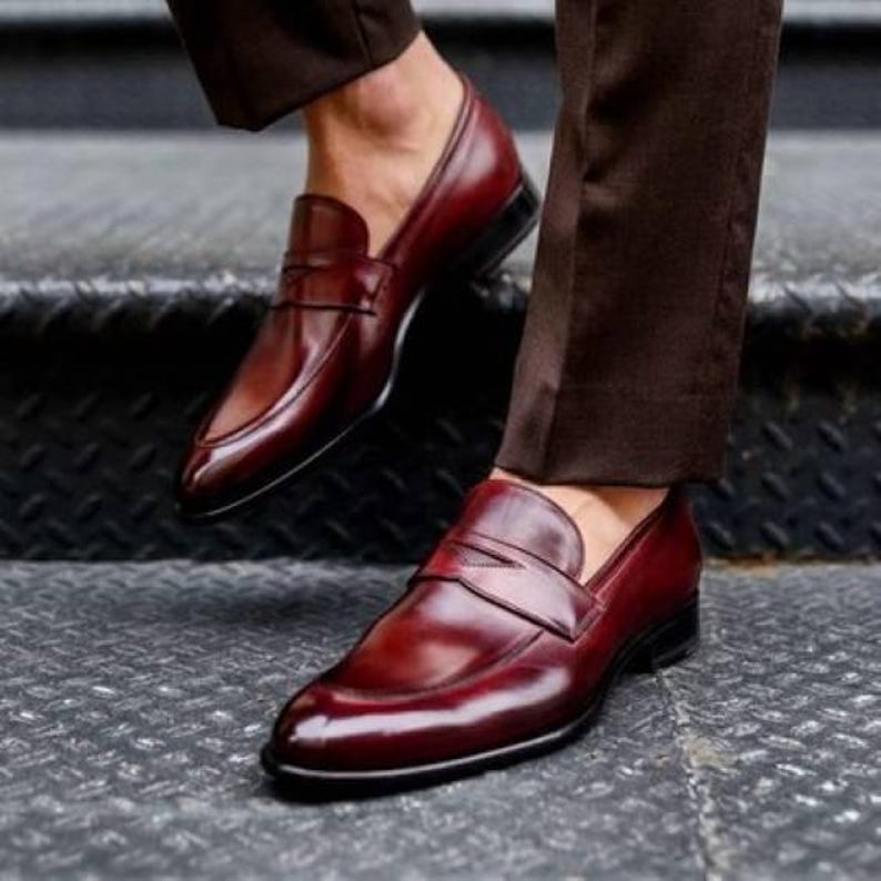 May include: Close-up of burgundy leather loafers with a classic design. The shoes have a polished finish and a slip-on style. The image shows the shoes on a textured, dark surface.