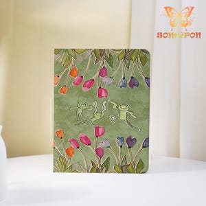 Op de afbeelding: Een rechthoekig notitieboek met een groene achtergrond en een speelse illustratie van drie lichtgroene kikkers die dansen tussen kleurrijke tulpen in tinten roze, oranje, paars en wit. In de rechterbovenhoek staan een vlinderlogo en de tekst somypon.