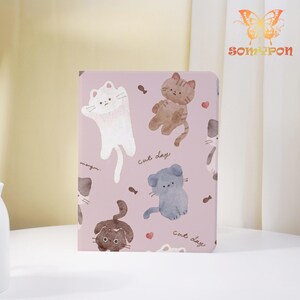 May include: A pink notebook with a repeating pattern of cartoon cats, fish, and hearts. The notebook has the words "magic" and "cat day" written on it. The notebook is a cute and whimsical item.