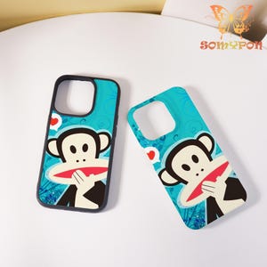 May include: Two phone cases featuring a cartoon monkey design. The cases have a teal background and a black border. The monkey has a white face, black ears, and a red mouth. Small red hearts are also visible.