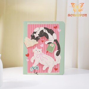 May include: A green and pink striped notebook with illustrations of two cats, books, hearts, and a game controller. One cat is white with a pink bow, and the other is black and white. The notebook has the brand name "SOMYPON" in the upper right corner.