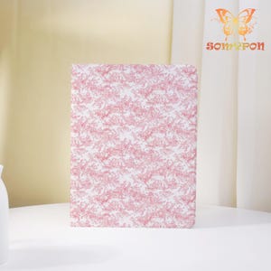 May include: A rectangular notebook with a pink and white floral pattern. The notebook is standing upright on a white surface. The notebook has rounded corners. The background is a light yellow and white.