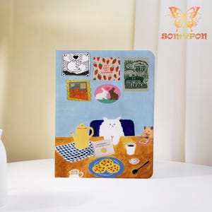 May include: A whimsical notebook with a blue cover featuring a cat illustration at a table with cookies, a teapot, and coffee. The cover also has several small sticker-like images with various designs and text. The notebook has a cute and playful theme.