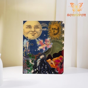 May include: A notebook with a colorful illustration featuring a smiling moon, a sun, and a woman with long black hair. The artwork includes fairies, flowers, and stars, set against a dark blue background. The notebook has a decorative design.