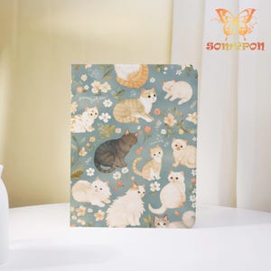 May include: A notebook with a teal background featuring illustrations of various cats and floral patterns. The cats are in different poses and colors, including gray, orange, and white. The notebook has a white border and is on a white surface.