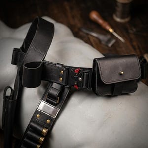 May include: Black leather utility belt with a holster, pouch, and cartridge holders. The belt features a metal buckle with a red release button and brass rivets. The belt is designed for carrying various items.