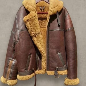 Puede incluir: Chaqueta de cuero marrón con cuello y forro de borrego. La chaqueta tiene cierre de cremallera, bolsillos y detalles de hebilla. El texto "IYALEATHER" es visible en la parte inferior.