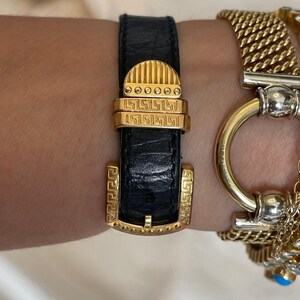 May include: Black leather watch strap with gold-tone buckle and accents. The buckle features a rectangular shape with a Greek key design. A gold chain bracelet is visible in the background.