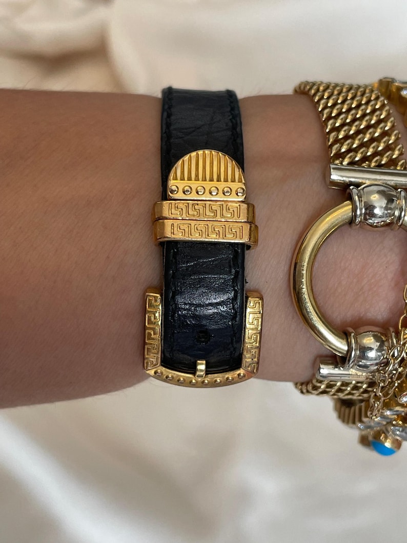 May include: Black leather watch strap with gold-tone buckle and accents. The buckle features a rectangular shape with a Greek key design. A gold chain bracelet is visible in the background.