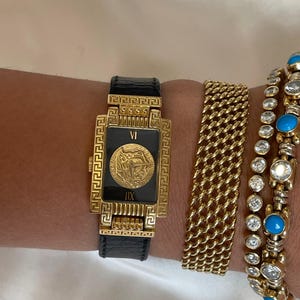 May include: A gold-toned rectangular watch with a black face and a Medusa head emblem, paired with gold bracelets. The watch has a black leather band. Additional bracelets include a gold chain link and a gold bracelet with turquoise and clear stones.