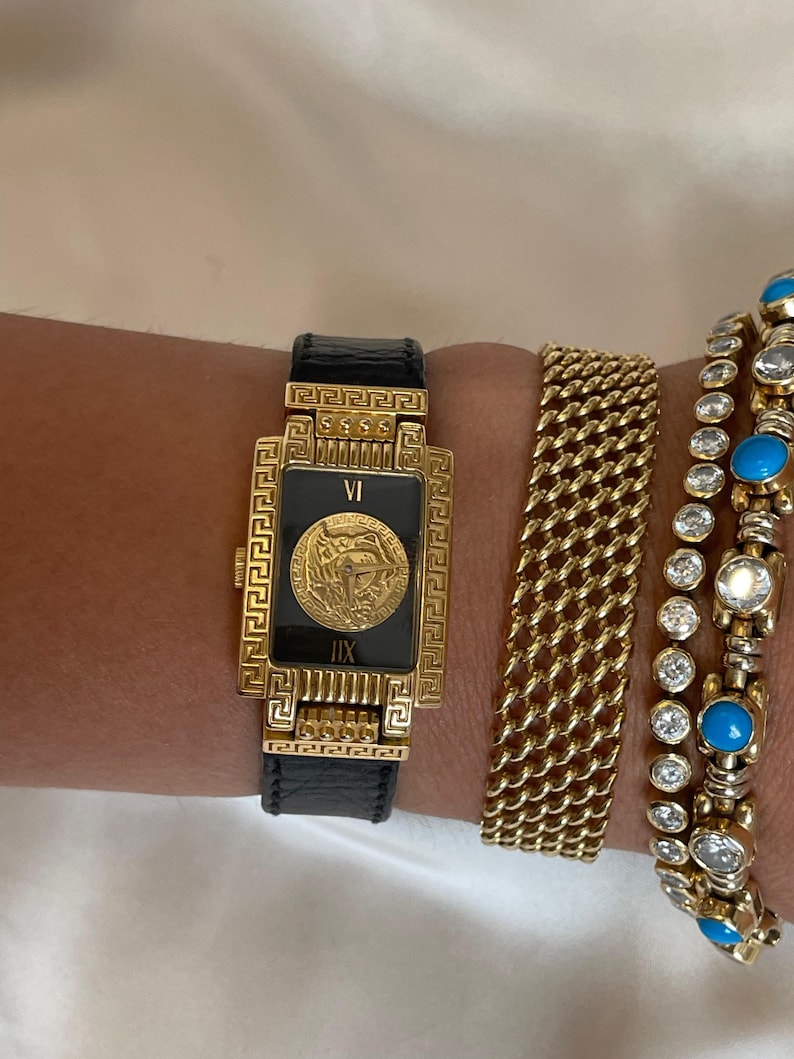May include: A gold-toned rectangular watch with a black face and a Medusa head emblem, paired with gold bracelets. The watch has a black leather band. Additional bracelets include a gold chain link and a gold bracelet with turquoise and clear stones.