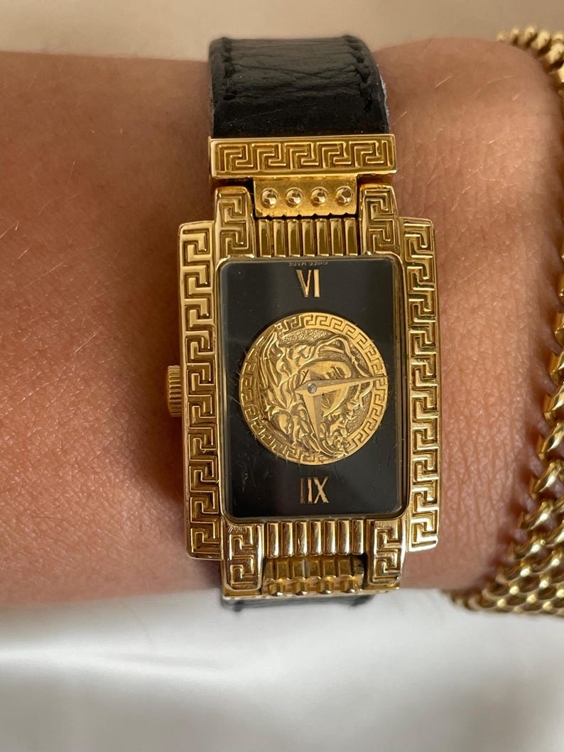 May include: Gold-toned rectangular watch with a black face and a black leather band. The watch face features Roman numerals and a gold medallion. The watch has a gold chain bracelet next to it.