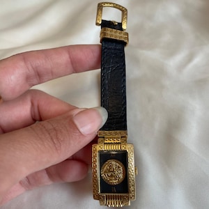 May include: A gold-toned rectangular wristwatch with a black face and a black leather band. The watch face features a gold emblem and Roman numerals. The watch has a gold buckle and a textured leather band.