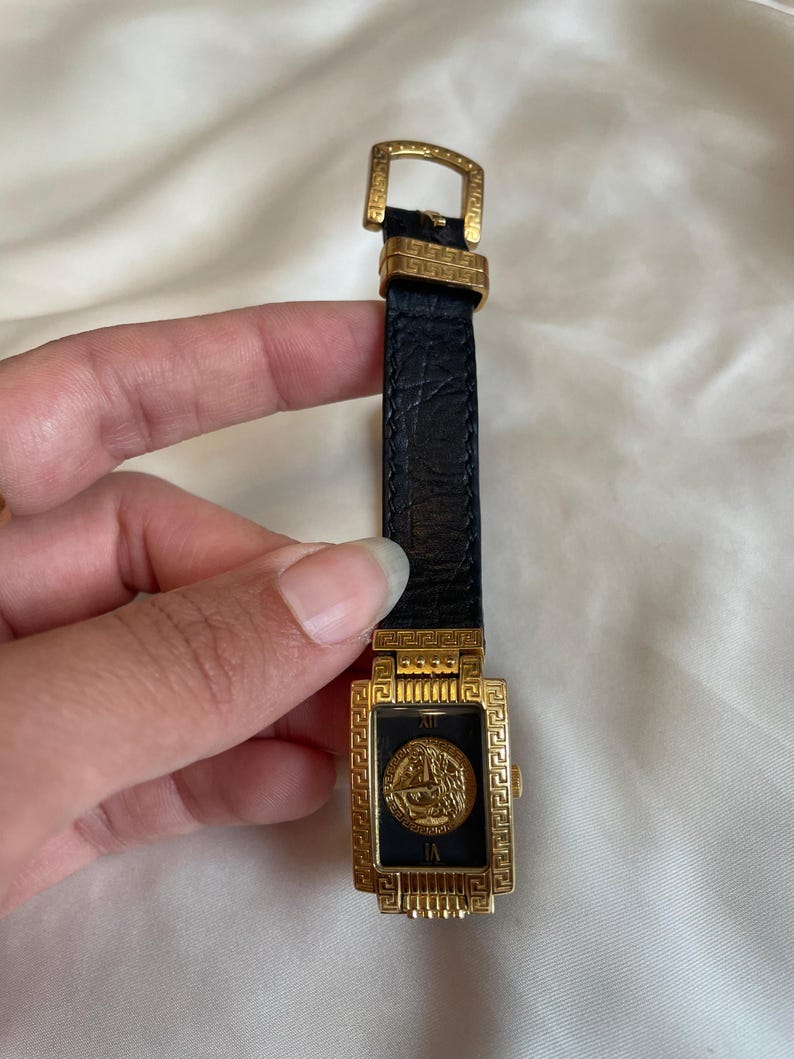 May include: A gold-toned rectangular wristwatch with a black face and a black leather band. The watch face features a gold emblem and Roman numerals. The watch has a gold buckle and a textured leather band.