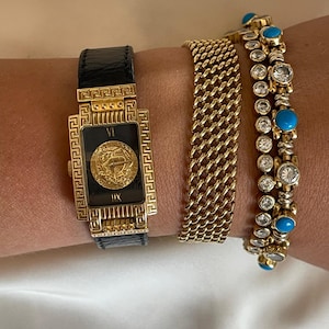 May include: A collection of gold jewelry, including a rectangular watch with a black leather strap and a gold face with Roman numerals. Two gold bracelets, one with a chain link design and another with turquoise and clear stones, complete the set.