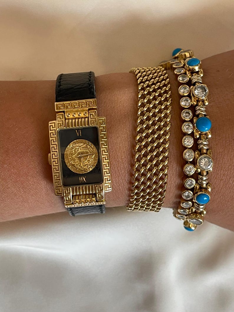 May include: A collection of gold jewelry, including a rectangular watch with a black leather strap and a gold face with Roman numerals. Two gold bracelets, one with a chain link design and another with turquoise and clear stones, complete the set.