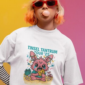 May include: White t-shirt with a graphic of a crying pink bunny holding a cookie, surrounded by Christmas lights and presents. The text "Tinsel Tantrum Tour '25" is above the bunny. The person is wearing red sunglasses and blowing a bubble.