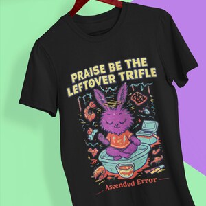May include: Black t-shirt with a colorful graphic of a purple rabbit meditating in a bowl, with the text "Praise Be The Leftover Trifle" and "Ascended Error". The rabbit wears a purple hat and orange shirt.