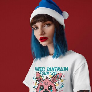 May include: White t-shirt with a cartoon graphic of a crying pink bunny holding a cookie, with the text "Tinsel Tantrum Tour '25". The model is wearing a blue and white Santa hat. The background is a solid red.