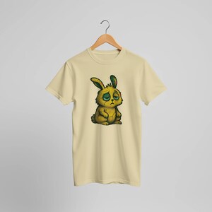 May include: A light yellow t-shirt featuring a cartoon illustration of a sad-looking yellow bunny with green accents. The bunny has droopy eyes and a distressed expression. The t-shirt is hanging on a wooden hanger.