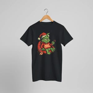 May include: Black t-shirt featuring a grumpy green cartoon character wearing a Santa hat and a red outfit with the text "MERRY WHATEVER". The character holds a candy cane and sits on a red sack with a small gift.