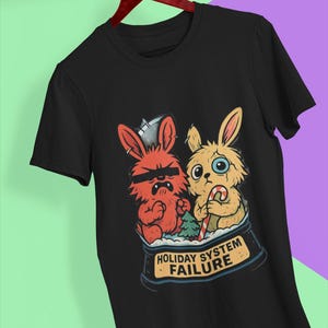 Może przedstawiać: Czarna koszulka z kreskówkową grafiką przedstawiającą dwa króliki wewnątrz kuli śnieżnej. Jeden czerwony królik nosi okulary przeciwsłoneczne i wygląda na złego, a drugi, żółty królik, wygląda na smutnego i trzyma laskę cukrową. Poniżej znajduje się napis "HOLIDAY SYSTEM FAILURE".