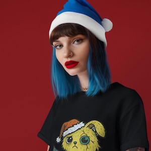 Puede incluir: Una camiseta negra con un conejito amarillo de dibujos animados con un gorro de Papá Noel y un pequeño árbol de Navidad. La persona de la imagen lleva un gorro de Papá Noel azul y blanco y tiene el pelo azul.