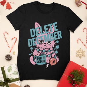 May include: Black t-shirt with a pink bunny graphic and the text "DELETE DECEMBER" in blue. The bunny is holding a mug that says "DELETE DECEMBER" and a Christmas ornament. Christmas-themed gifts and decorations surround the shirt.
