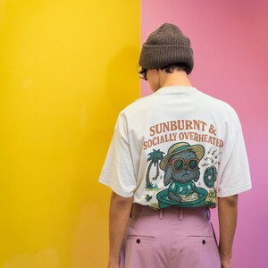 May include: A person wearing a white t-shirt with the text "SUNBURNT & SOCIALLY OVERHEATED" and a graphic of a dog in sunglasses. The person is wearing a brown beanie and pink pants. The background is yellow and pink.