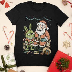 May include: Black t-shirt featuring a cartoon illustration of Santa Claus and a green character, both with grumpy expressions. The image includes a candy cane, gifts, and the text "Merry Christmas". Festive holiday apparel.