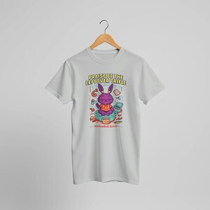 May include: A light gray t-shirt featuring a cartoon illustration of a purple rabbit in a bowl, surrounded by food items. The text "Praise Be The Leftover Trifle" and "Ascended Error" are printed on the shirt.