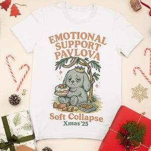 May include: White t-shirt with the text "EMOTIONAL SUPPORT PAVLOVA" and a cartoon dog holding a cake. The shirt also has the text "Soft Collapse Xmas'25". Christmas-themed gifts and decorations surround the shirt.