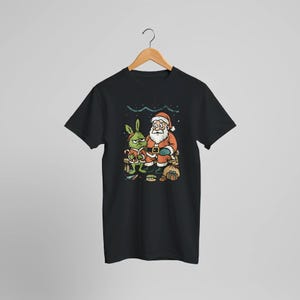 May include: Black t-shirt featuring a cartoon illustration of Santa Claus and a green creature. Santa is wearing a red suit and hat, while the green creature is holding a candy cane. The design includes gifts and food items, with a festive, humorous theme.