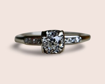 May include: A silver ring with a large, clear, square-cut diamond in the center. Smaller diamonds are set on either side of the center stone.