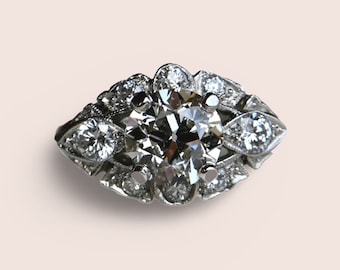 Puede incluir: Un anillo de plata con un gran diamante redondo y claro rodeado de diamantes más pequeños. El anillo tiene un diseño floral delicado.
