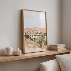 May include: A framed landscape painting with a light wood frame. The artwork depicts a desert scene with peach-colored mountains, green foliage, and a pale blue sky. Two white vases and a stack of books sit on a wooden shelf.