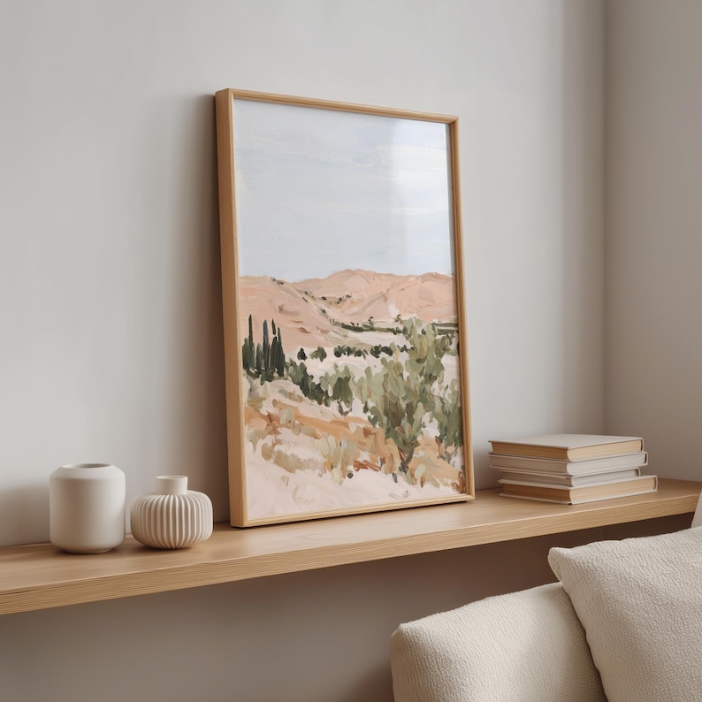 May include: A framed landscape painting with a light wood frame. The artwork depicts a desert scene with peach-colored mountains, green foliage, and a pale blue sky. Two white vases and a stack of books sit on a wooden shelf.