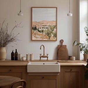 May include: A kitchen scene featuring a framed landscape painting with a light blue sky and rolling hills. A white farmhouse sink with a brass faucet is centered below the painting. Wooden countertops and cabinets complement the neutral color palette.