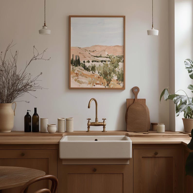 May include: A kitchen scene featuring a framed landscape painting with a light blue sky and rolling hills. A white farmhouse sink with a brass faucet is centered below the painting. Wooden countertops and cabinets complement the neutral color palette.