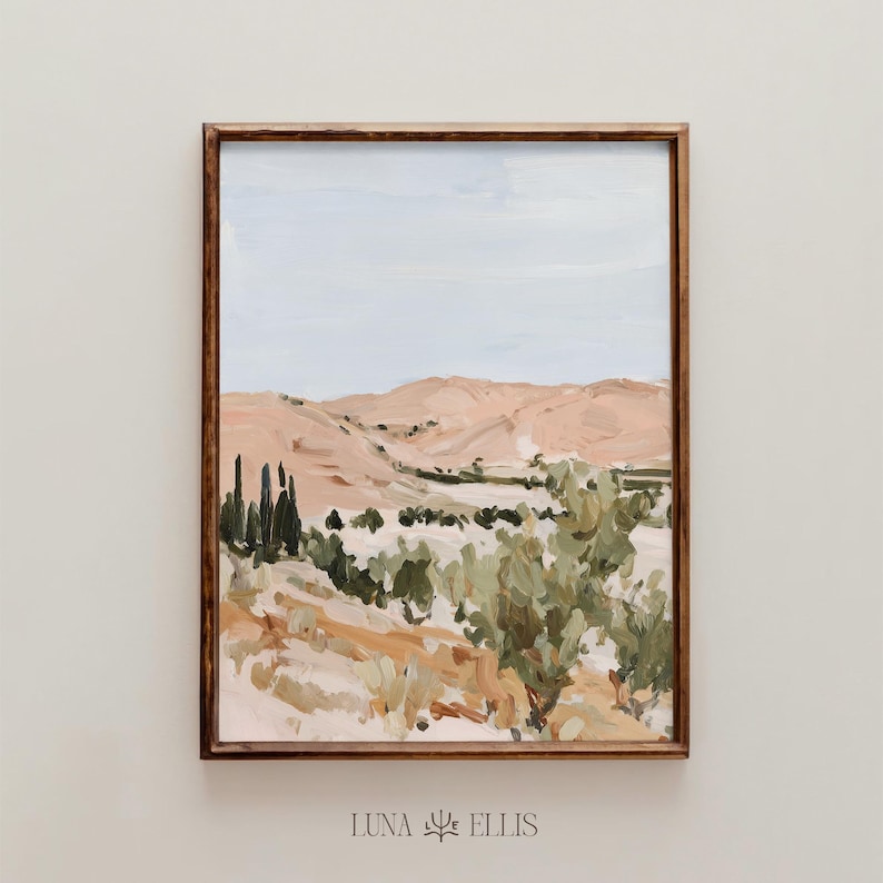 May include: A framed landscape painting featuring a desert scene with rolling hills in shades of peach and tan, under a pale blue sky. Green foliage and tall, dark green trees are in the foreground. The frame is a dark brown color. The text "LUNA ELLIS" is in the bottom right corner.