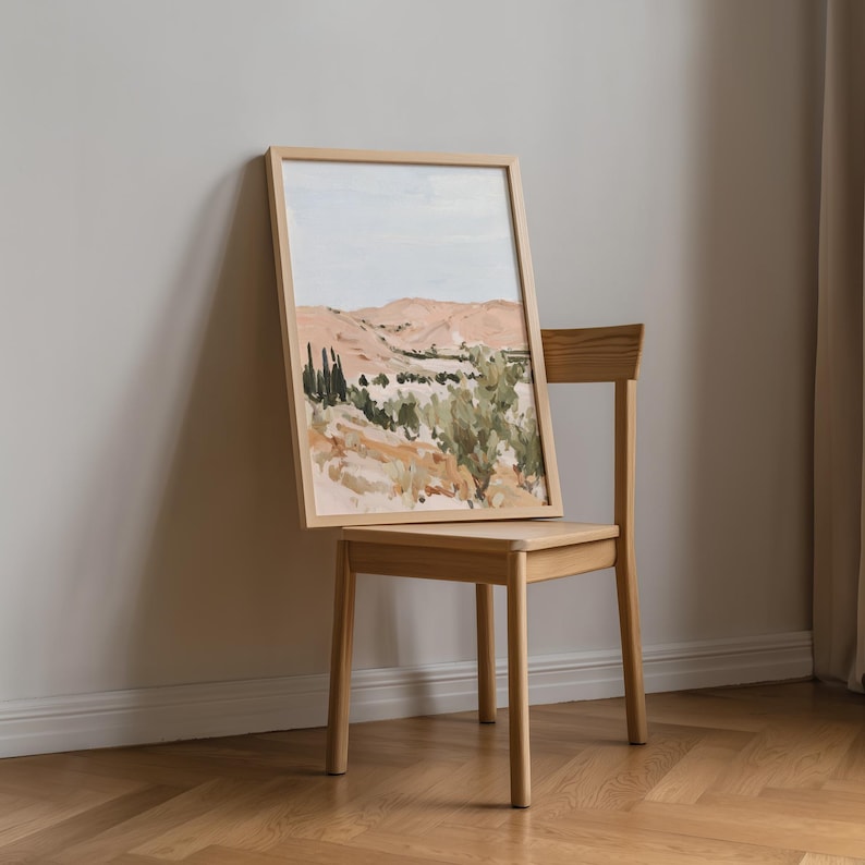 May include: A framed landscape painting depicting rolling hills in shades of beige, green, and blue. The artwork is displayed on a light-colored wooden chair, leaning against a wall. The painting's frame is also light-colored wood.