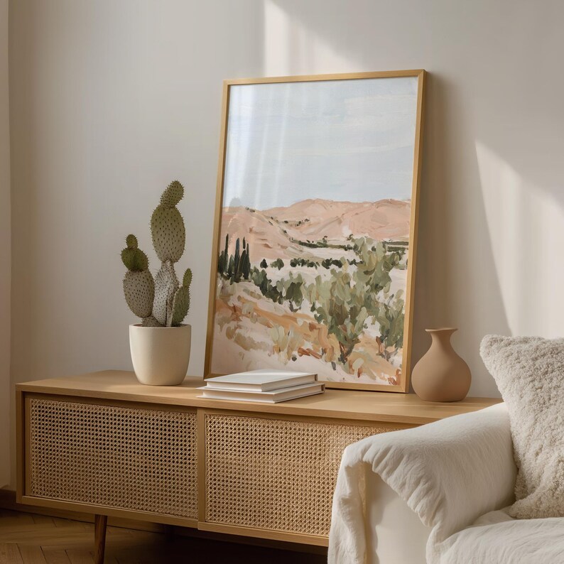 May include: A framed landscape art print with a light wood frame. The artwork depicts a desert scene with muted tones of peach, green, and blue. A potted cactus and a small vase sit on a wooden cabinet. A white sofa is in the foreground.
