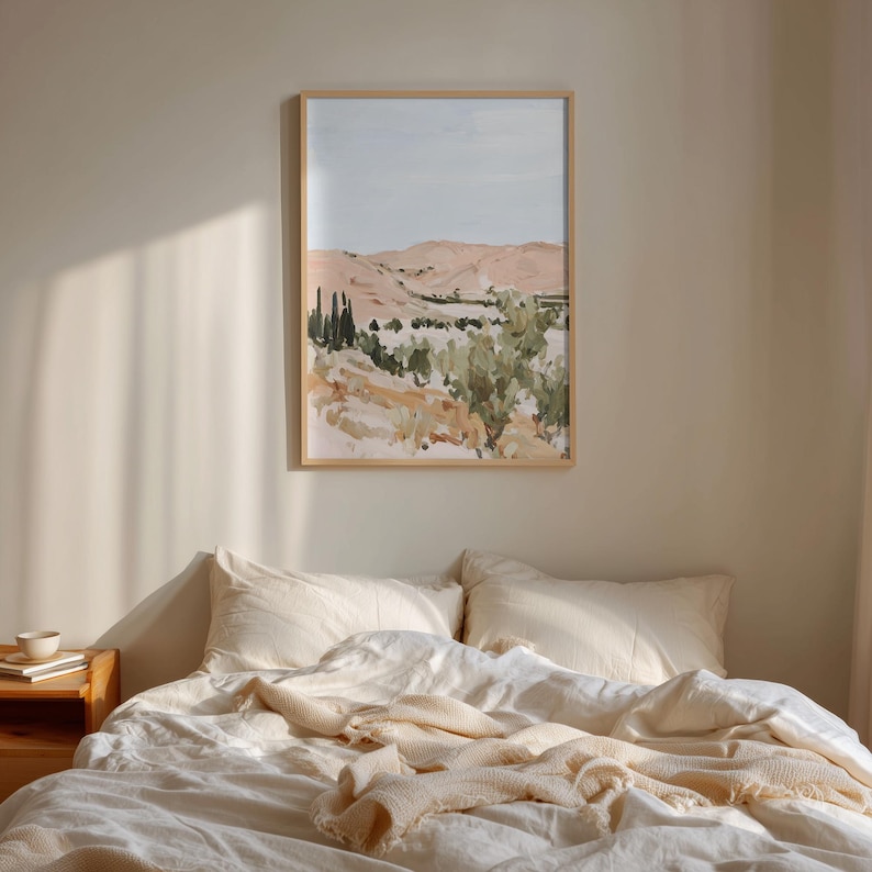 May include: A framed landscape painting with a light wood frame hangs above a bed. The artwork depicts a desert scene with peach-colored hills, green trees, and a pale blue sky. The bed has white linens and a cream-colored blanket.
