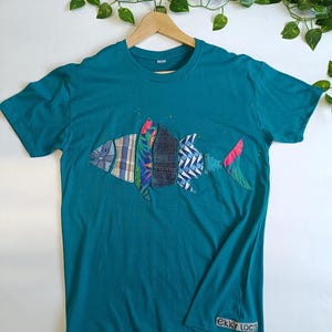 Puede incluir: Camiseta azul turquesa con un diseño de pez colorido hecho con recortes de tela. El pez está hecho de diferentes patrones y colores, incluyendo cuadros, flores y mezclilla. La camiseta tiene el texto "ekky loca" en la esquina inferior derecha.