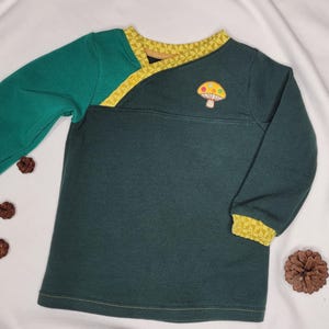 Könnte beinhalten: Ein langärmliges, dunkelgrünes Shirt mit einem kontrastierenden grünen Ärmel und gelben Besatz. Auf der Brust befindet sich ein Pilz-Applikation. Das Shirt ist mit Tannenzapfen verziert.