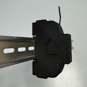 May include: Black plastic electrical component with a small wire sticking out. The component is mounted on a silver metal rail.