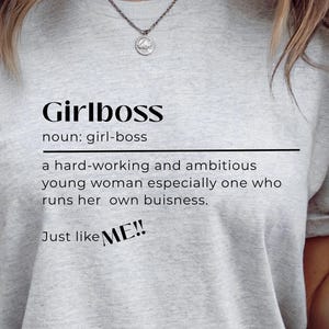 May include: A light gray t-shirt with the word "Girlboss" printed in black, along with a definition. The text "Just like ME!!" is also printed on the shirt. The shirt is made of a soft material.
