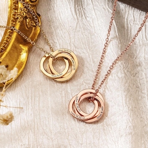 May include: Two personalised necklaces with interlocking rings. One is gold-toned, the other rose gold. Each ring is engraved with names and dates. The necklaces rest on a white, textured surface, with a gold-framed mirror in the background.