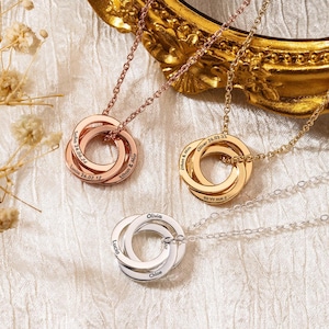 May include: Three personalised necklaces with interlocking rings in rose gold, gold, and silver. Each ring is engraved with names and dates. The necklaces are displayed on a textured white surface with a gold ornate frame in the background.