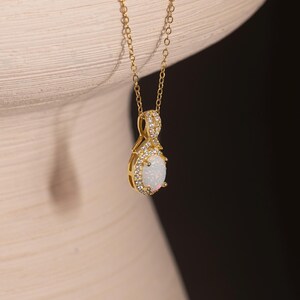 Peut inclure: Un collier doré composé d'une chaîne fine et d'un pendentif ovale. Le pendentif présente une pierre centrale blanche irisée entourée d'un halo de petites pierres claires, avec des pierres claires supplémentaires serties sur la bélière.